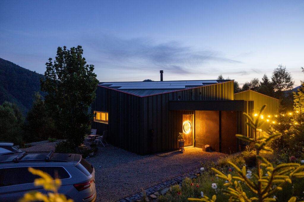 Exterior view of a cozy mountain cabin lit by warm string lights at dusk, surrounded by pine trees against a twilight sky