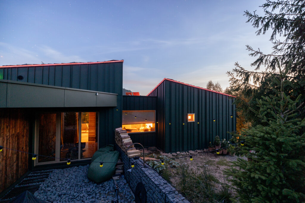 A cozy modern cabin under the evening sky, gently lit by garden string lights. The warm indoor glow contrasts beautifully with the twilight outdoors, creating a peaceful atmosphere
