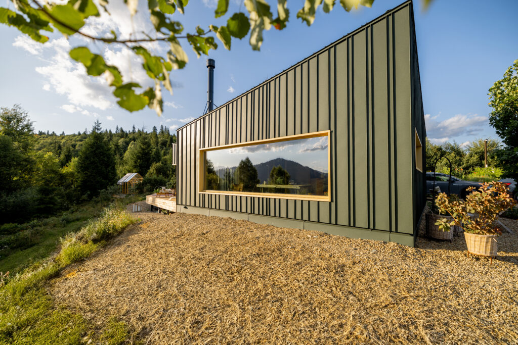 Modern green house with a large window reflecting a scenic mountain landscape. The house is set on a hillside, surrounded by natural vegetation under a clear sky, offering a peaceful rural retreat