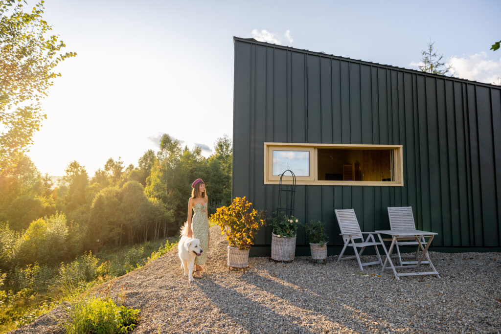 A woman walking with her white fluffy dog in front of a green modern house exterior, surrounded by nature during golden hour. Comfortable outdoor chairs and potted plants add to the serene setting