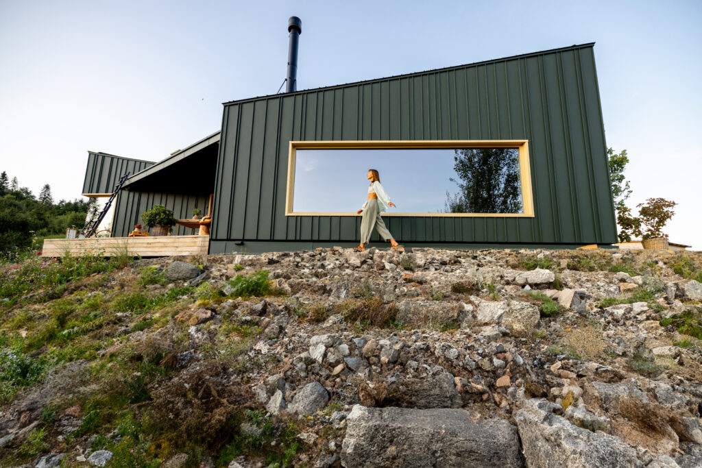 A woman stands calmly by a modern cabin with a large reflective window, embracing the tranquility of nature. The minimalist design and natural surroundings create a peaceful retreat