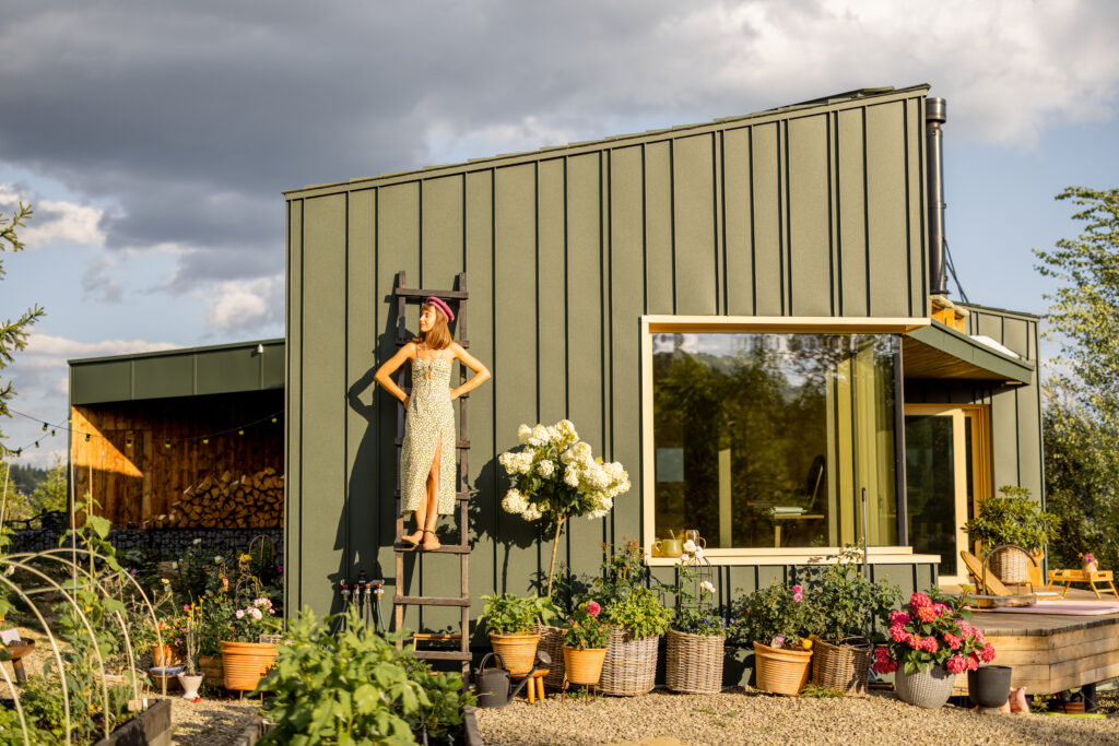 A woman in a summer dress stands on a ladder outside a modern garden house, surrounded by lush greenery and blooming flowers. The serene mountain backdrop adds to the tranquility of the scene
