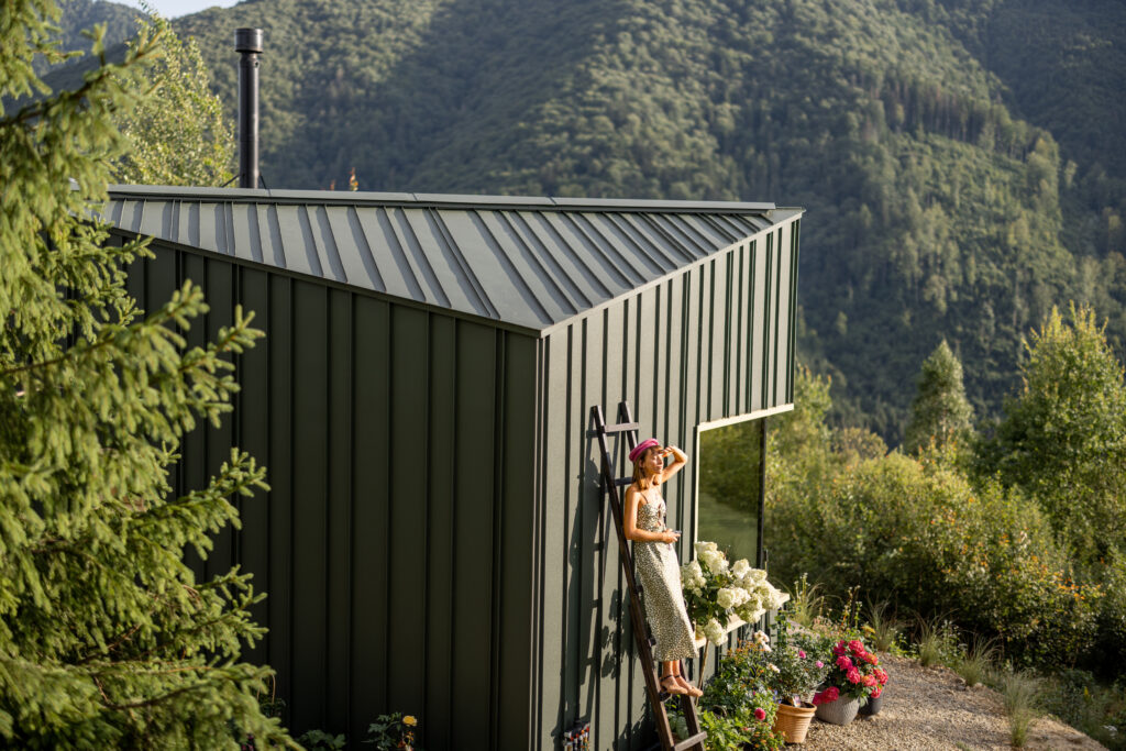 A woman stands on a ladder beside a modern eco-house surrounded by mountains and a blooming garden. The morning light adds warmth, highlighting the beauty of nature and the tranquility of rural life